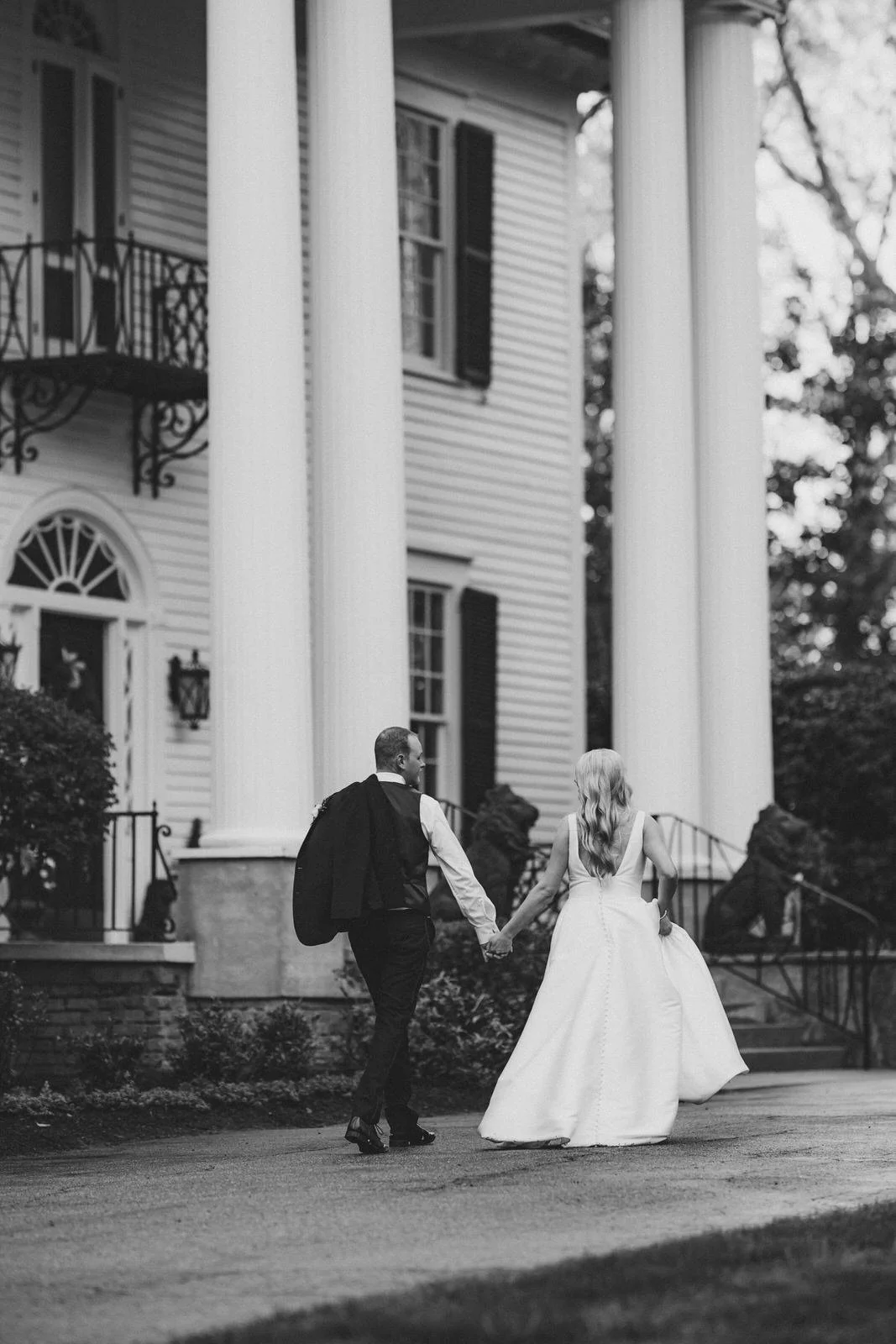 Black and white photo of a bride and groom walking hand in hand toward Duncan Estate’s front steps in Spartanburg, SC, with the groom’s jacket over his shoulder.
