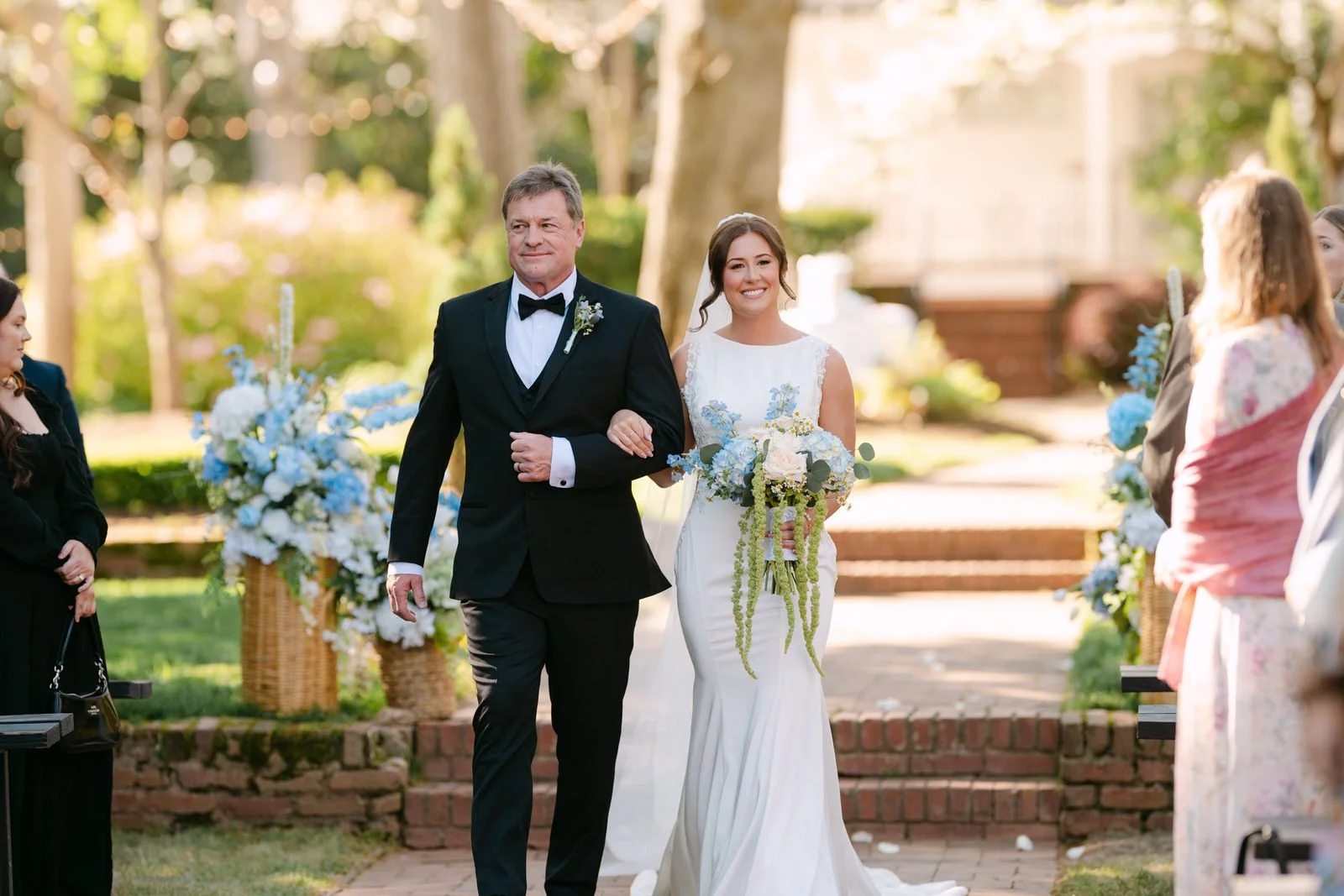 Smiling bride escorted down the brick aisle during a garden wedding ceremony at Duncan Estate in Spartanburg, SC, holding a cascading blue and white bouquet.