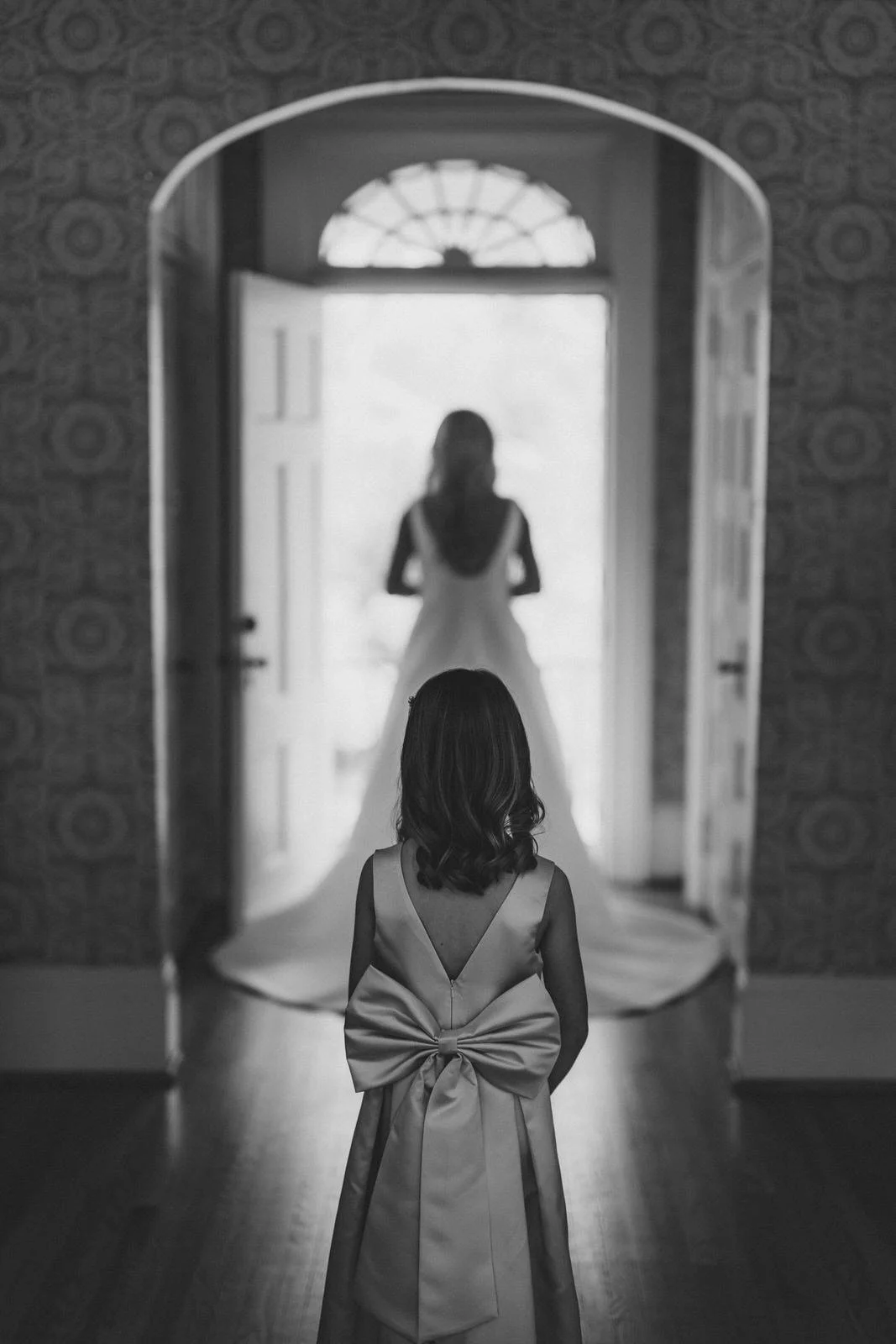 Black and white photo of a flower girl with a large bow on her dress looking toward the bride in Duncan Estate’s open doorway in Spartanburg, SC.