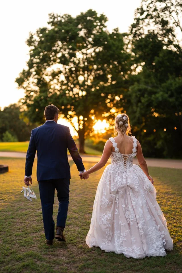 Bride and groom walking hand in hand across the lawn at Edinburgh West, with golden sunset light shining through the trees behind them.