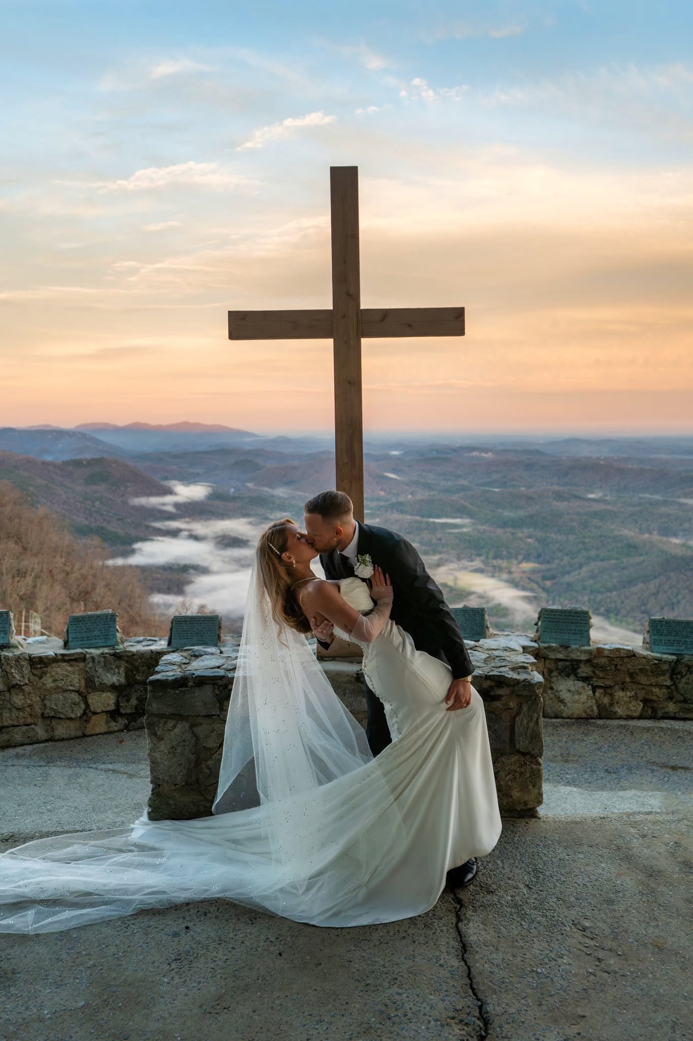 Groom dipping and kissing his bride beneath the wooden cross at Pretty Place Chapel, with her veil spread across the floor and pastel skies above misty mountains.