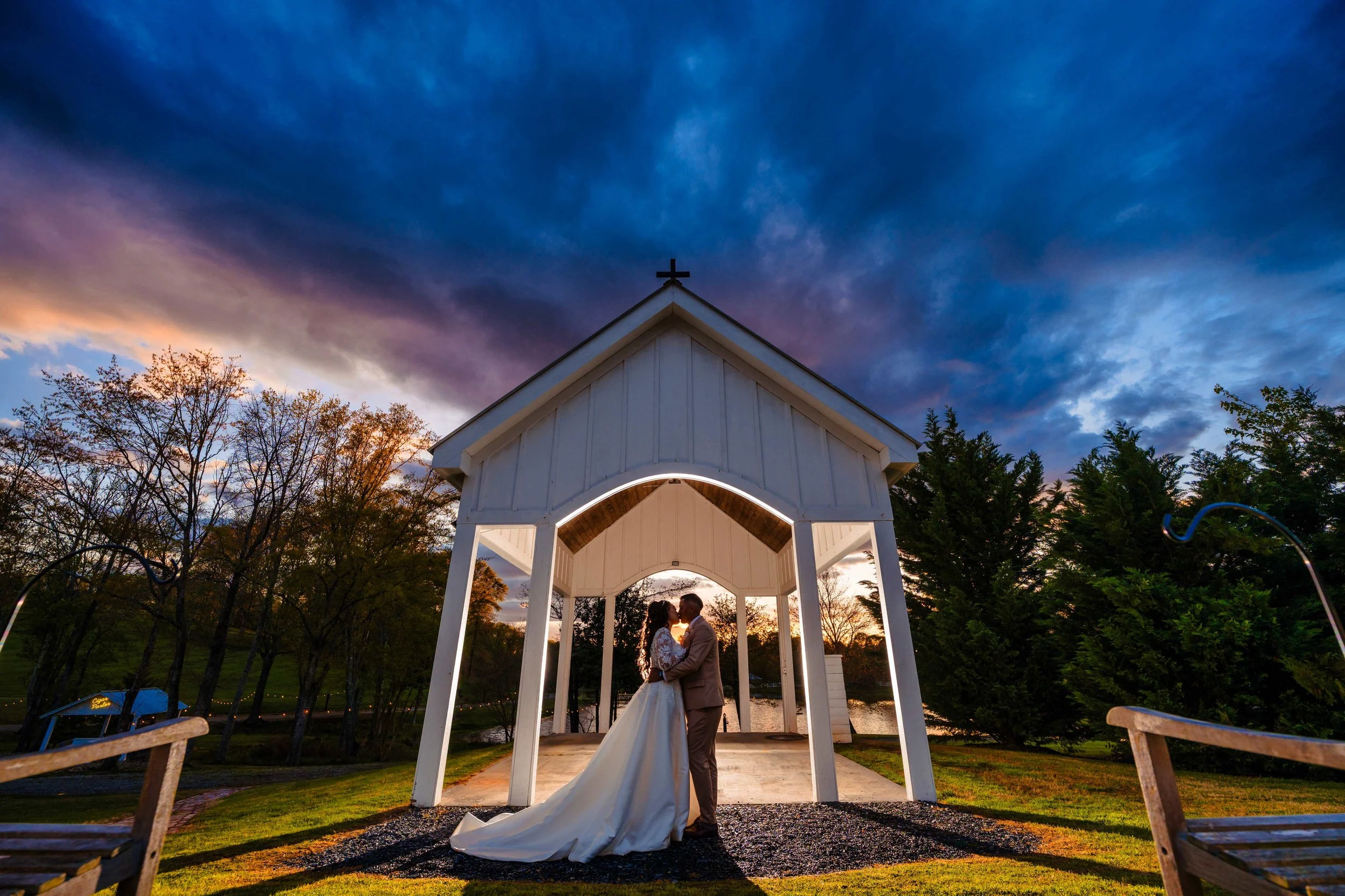 Bride and groom kissing beneath the white open-air chapel at South Wind Ranch, with blue and pink sunset clouds overhead and the pond behind them.