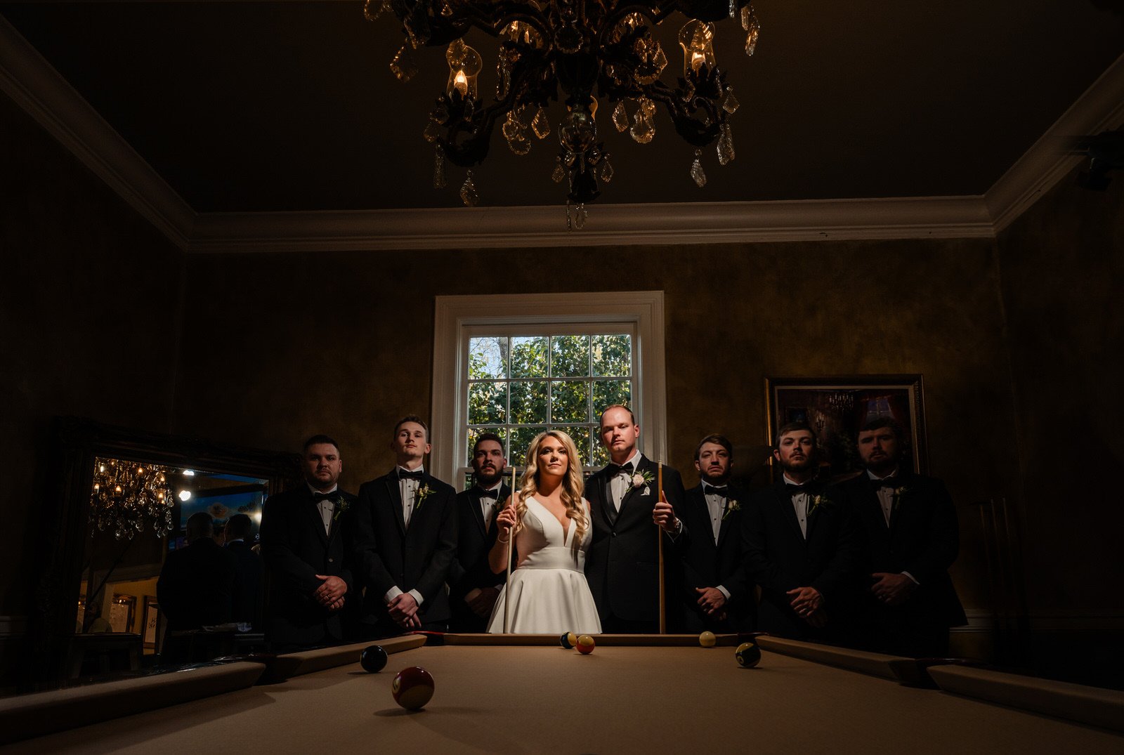Bride, groom, and six groomsmen posing behind a pool table beneath a chandelier inside Duncan Estate in Spartanburg, SC.
