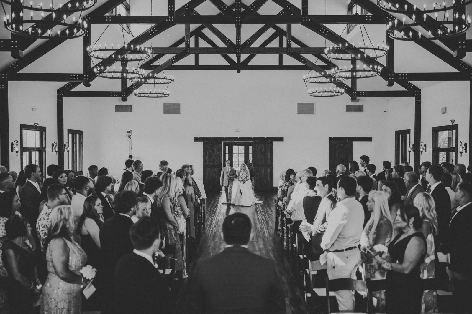 Black and white photo of a bride walking down the aisle with her escort at MeadowView, as guests stand beneath exposed beams and circular chandeliers.