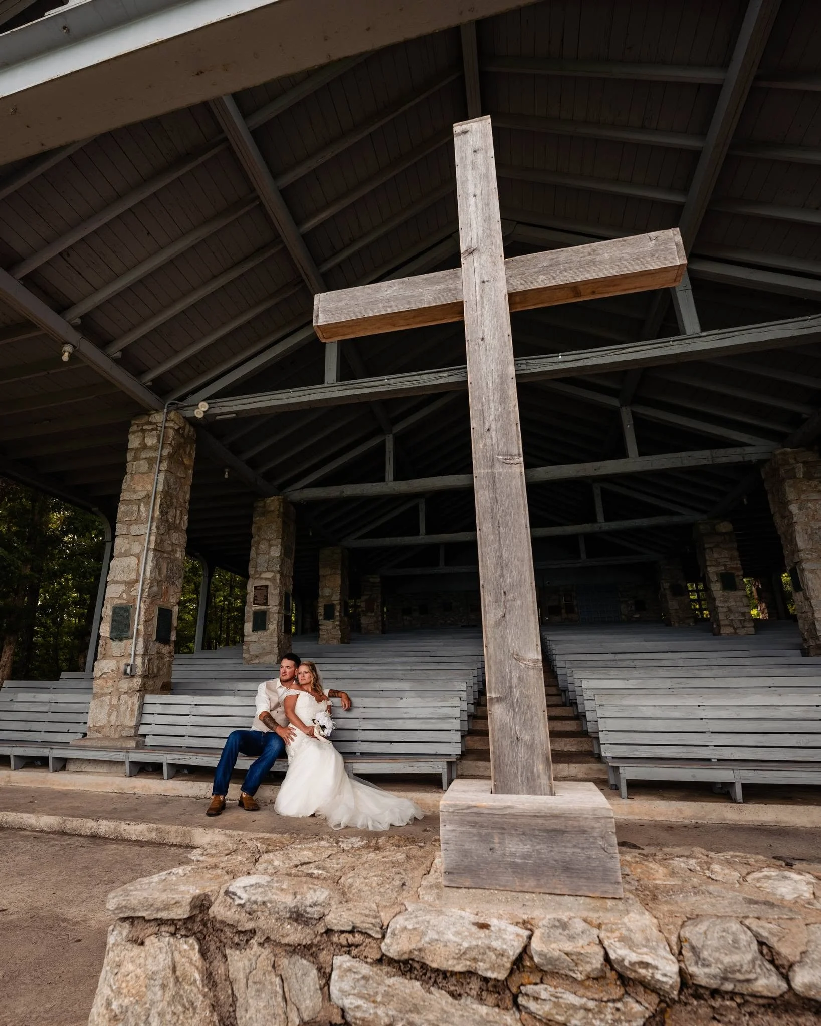 Bride and groom seated together on a wooden pew at Pretty Place Chapel, with a large wooden cross in the foreground and stone columns beneath exposed roof beams.