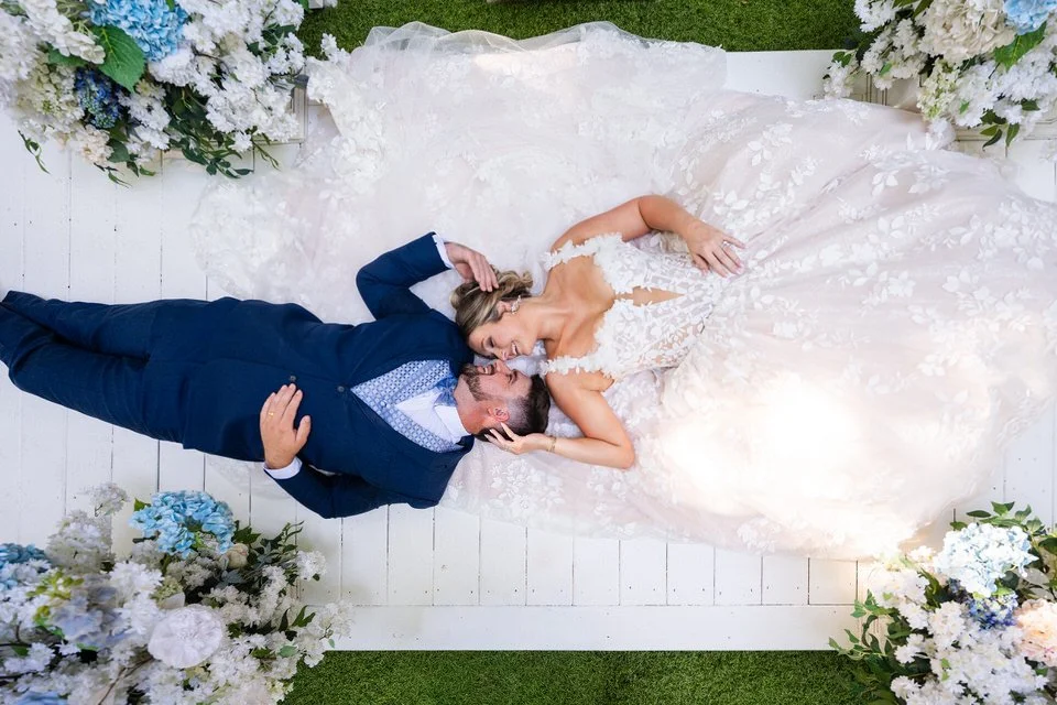 Overhead portrait of a bride and groom lying together on a white wooden platform at Edinburgh West, surrounded by blue and white flowers, with her lace gown spread around them.