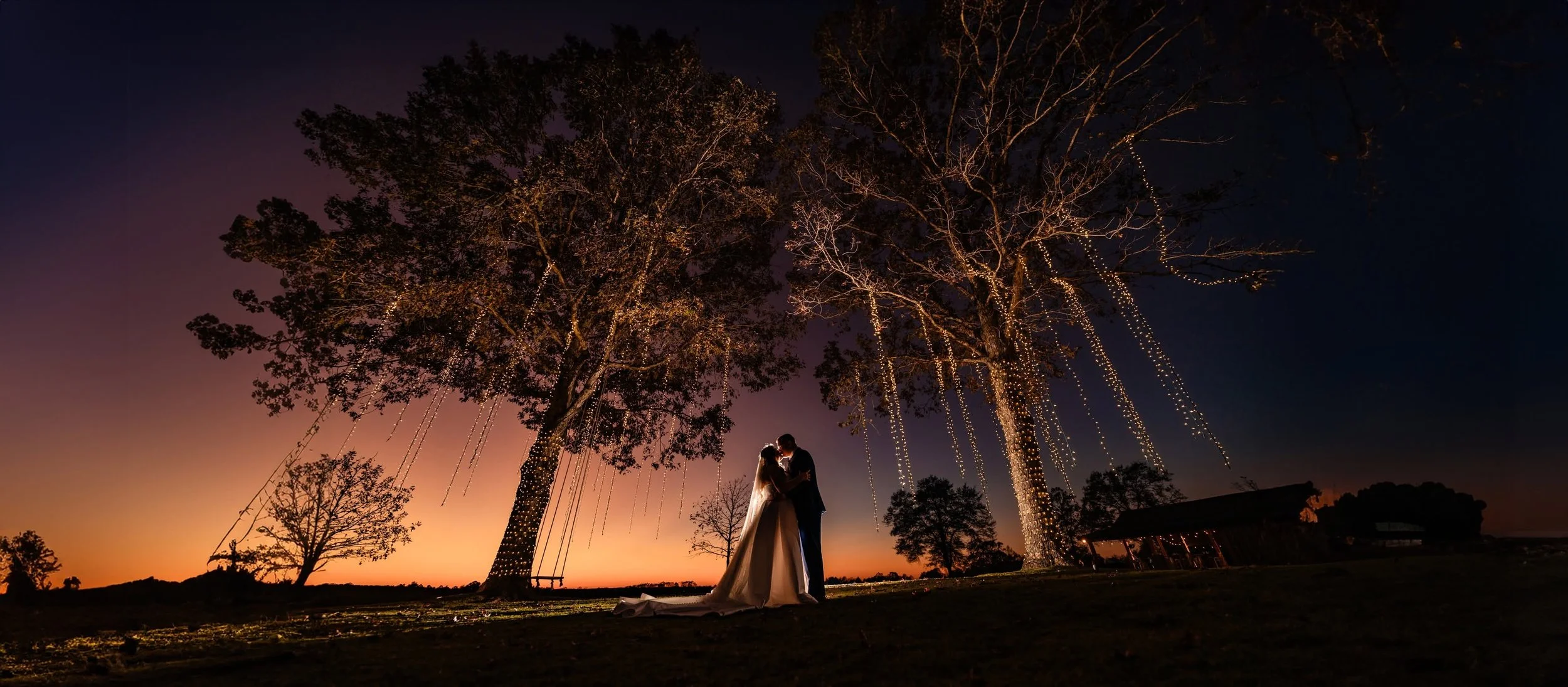 Bride and groom kissing beneath trees draped in string lights at Sunset Farm, against an orange and purple twilight sky.