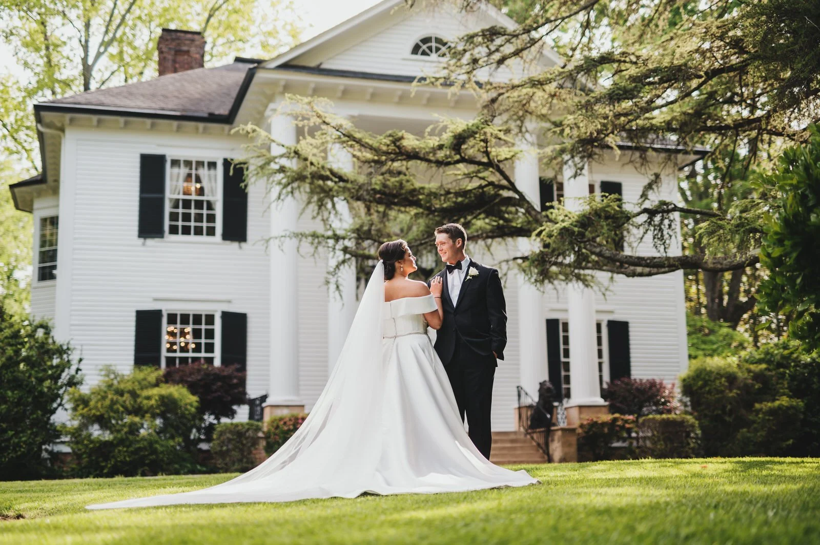 Bride and groom smiling at each other on the lawn at Duncan Estate, with the bride’s long train spread across the grass and the white historic house behind them.