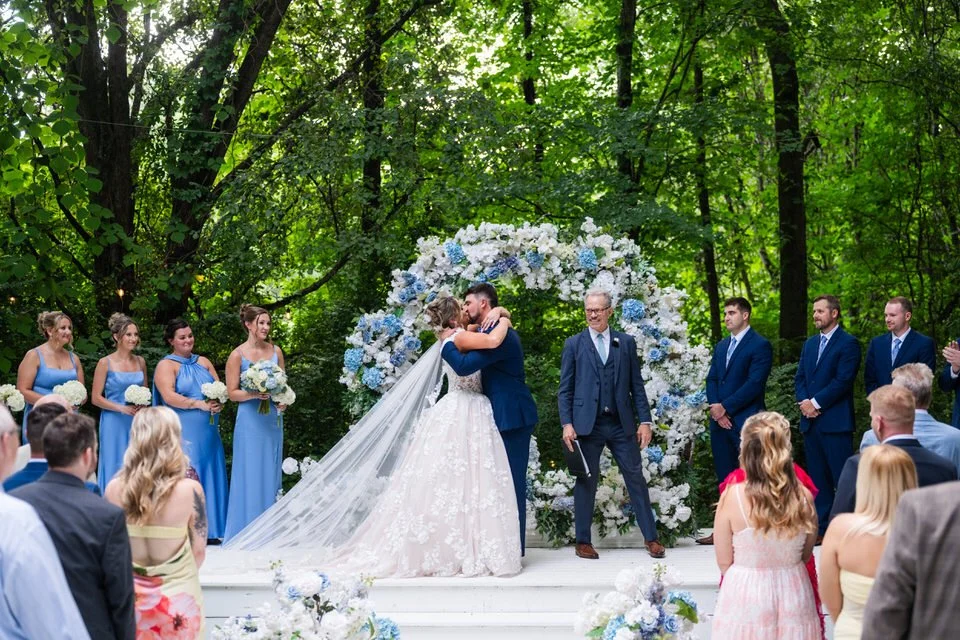 Bride and groom sharing their first kiss during an outdoor wedding ceremony at Edinburgh West, framed by a blue and white floral arch with their wedding party beside them.