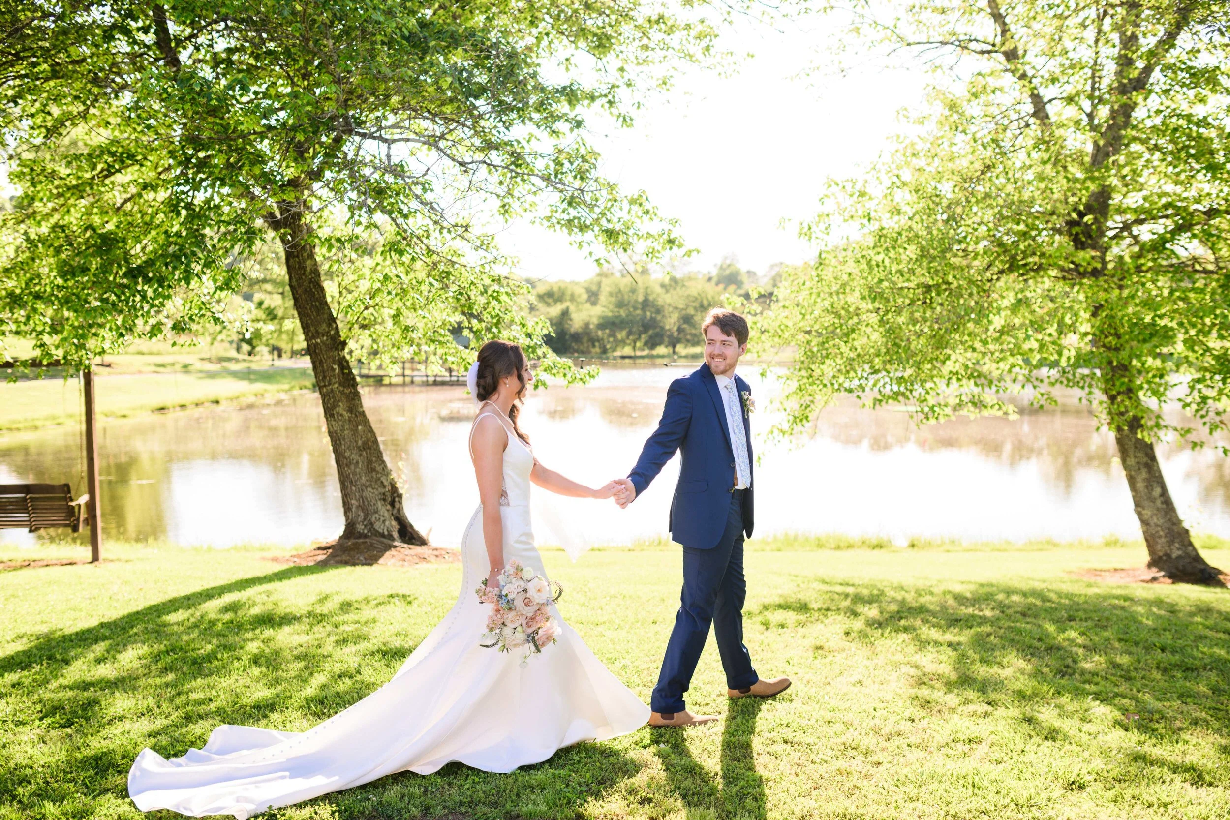 Bride and groom holding hands and smiling at each other while walking beneath leafy trees beside the pond at South Wind Ranch.