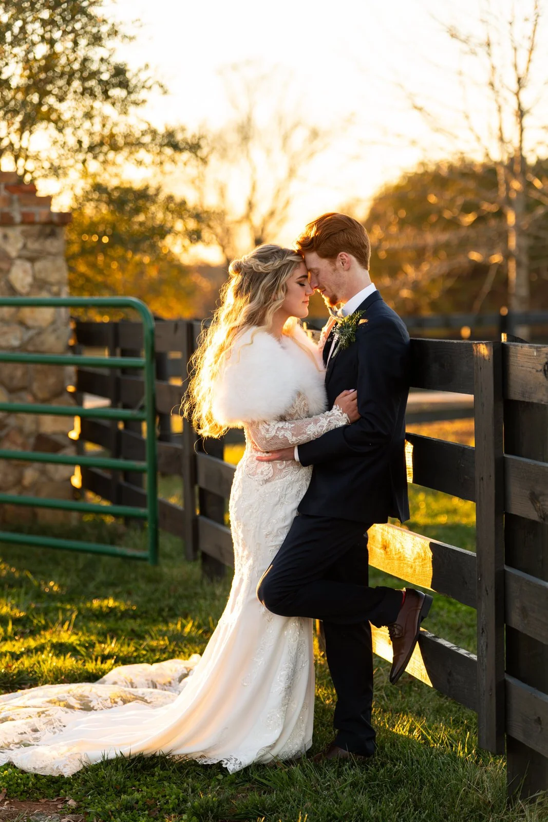Bride in a lace gown and white wrap embracing the groom beside MeadowView’s black pasture fence, with golden sunlight illuminating her hair.