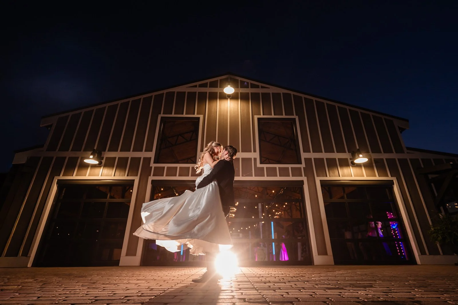Groom lifting and kissing his bride outside The Barn at Sitton Hill at night, with her dress sweeping outward and warm lights glowing behind them.