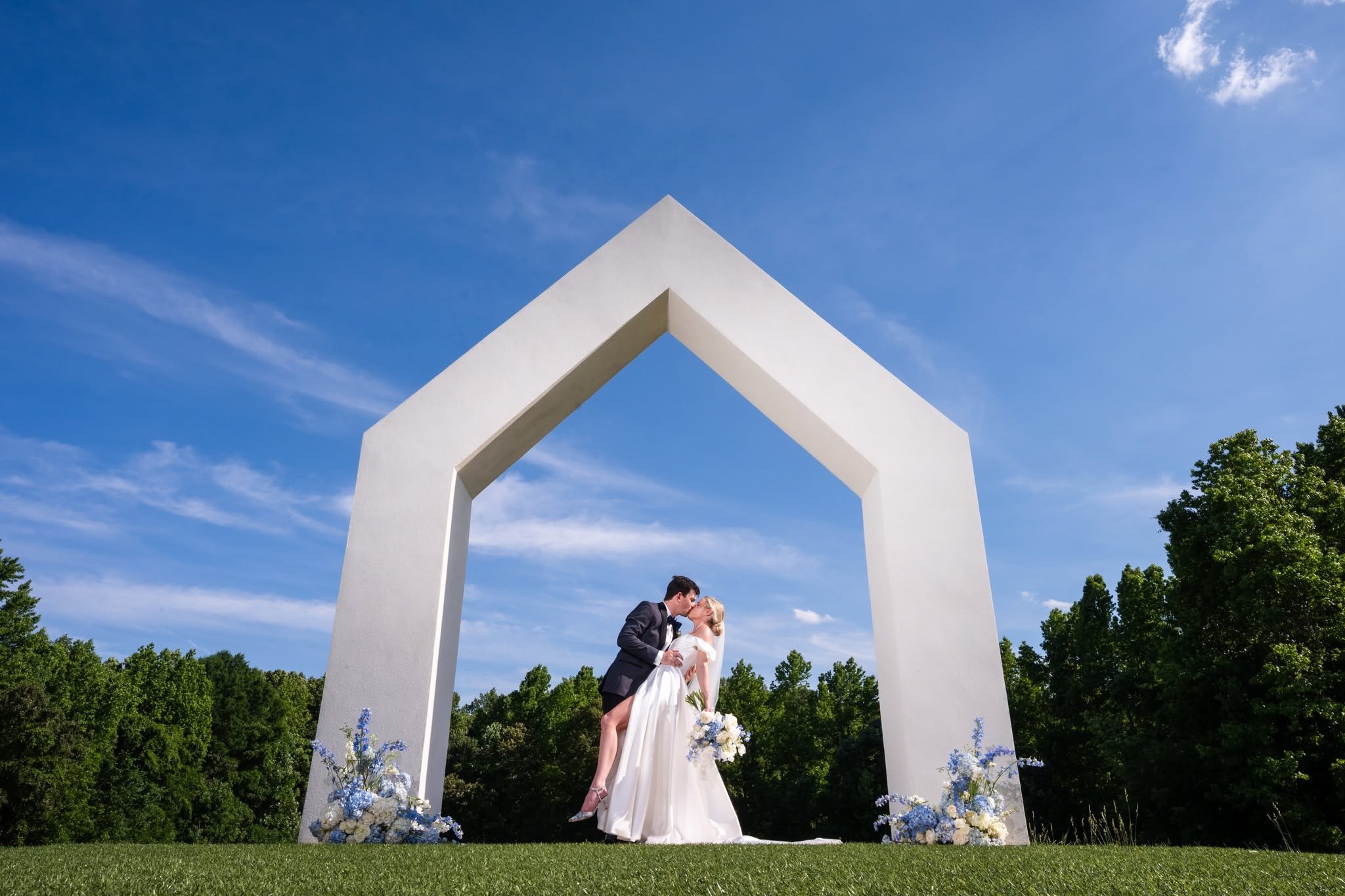 Bride and groom kissing beneath the large white ceremony arch at The Lumen House, framed by blue and white flowers and a bright blue sky.