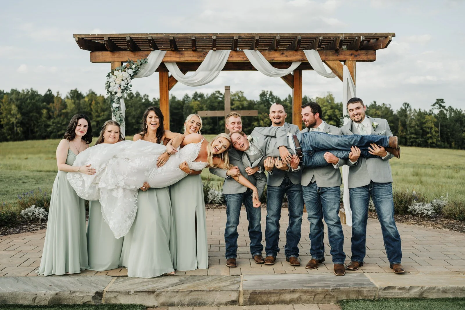 Bridesmaids in sage green dresses and groomsmen in gray jackets and jeans lifting the bride and groom in front of MeadowView’s wooden ceremony pergola.