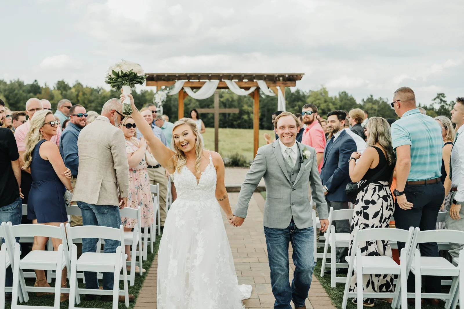 Smiling newlyweds holding hands as they walk down MeadowView’s outdoor ceremony aisle, with the bride raising her white bouquet and guests standing beside them.