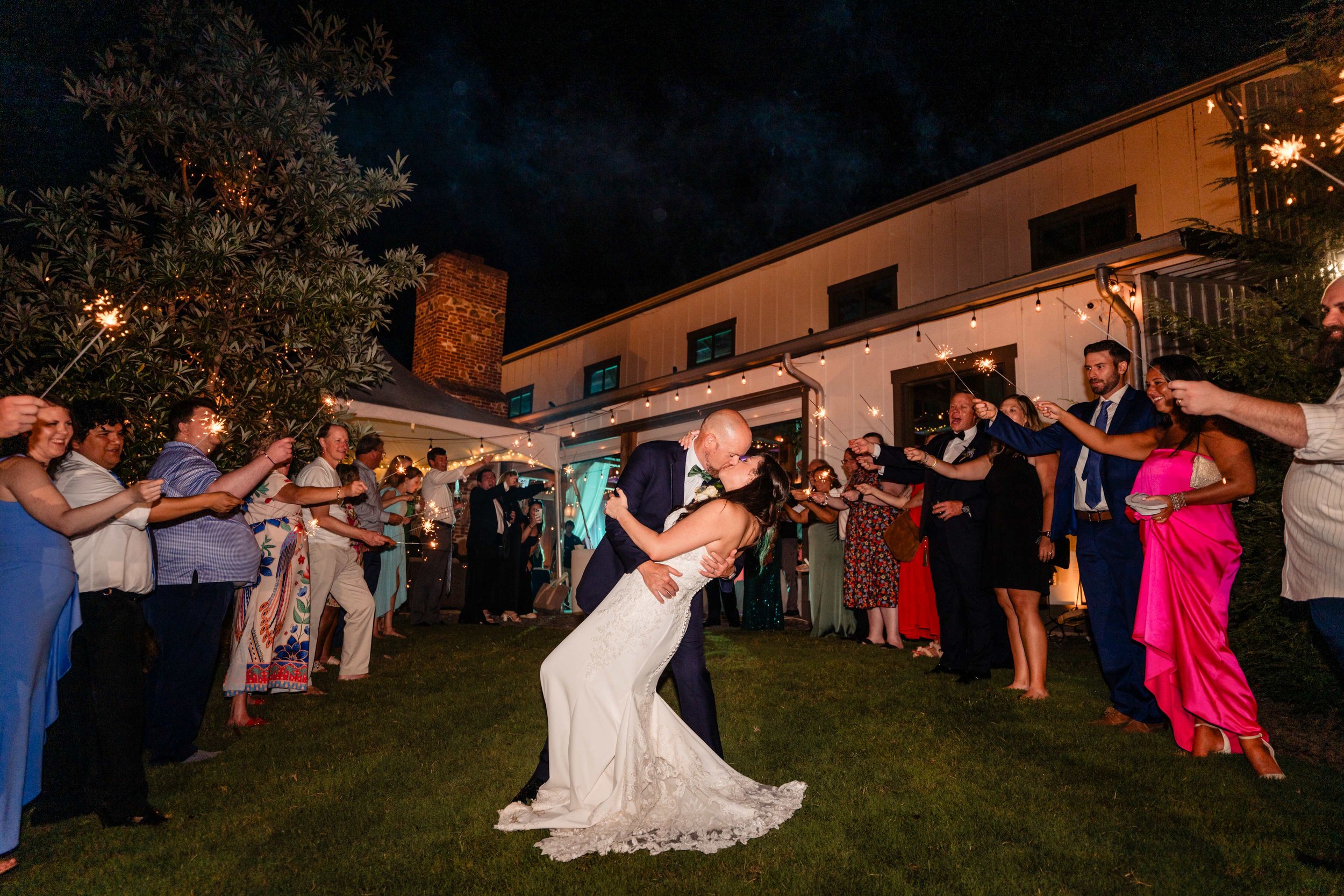 Groom dipping and kissing the bride during a nighttime sparkler exit at South Wind Ranch, surrounded by guests outside the reception barn.