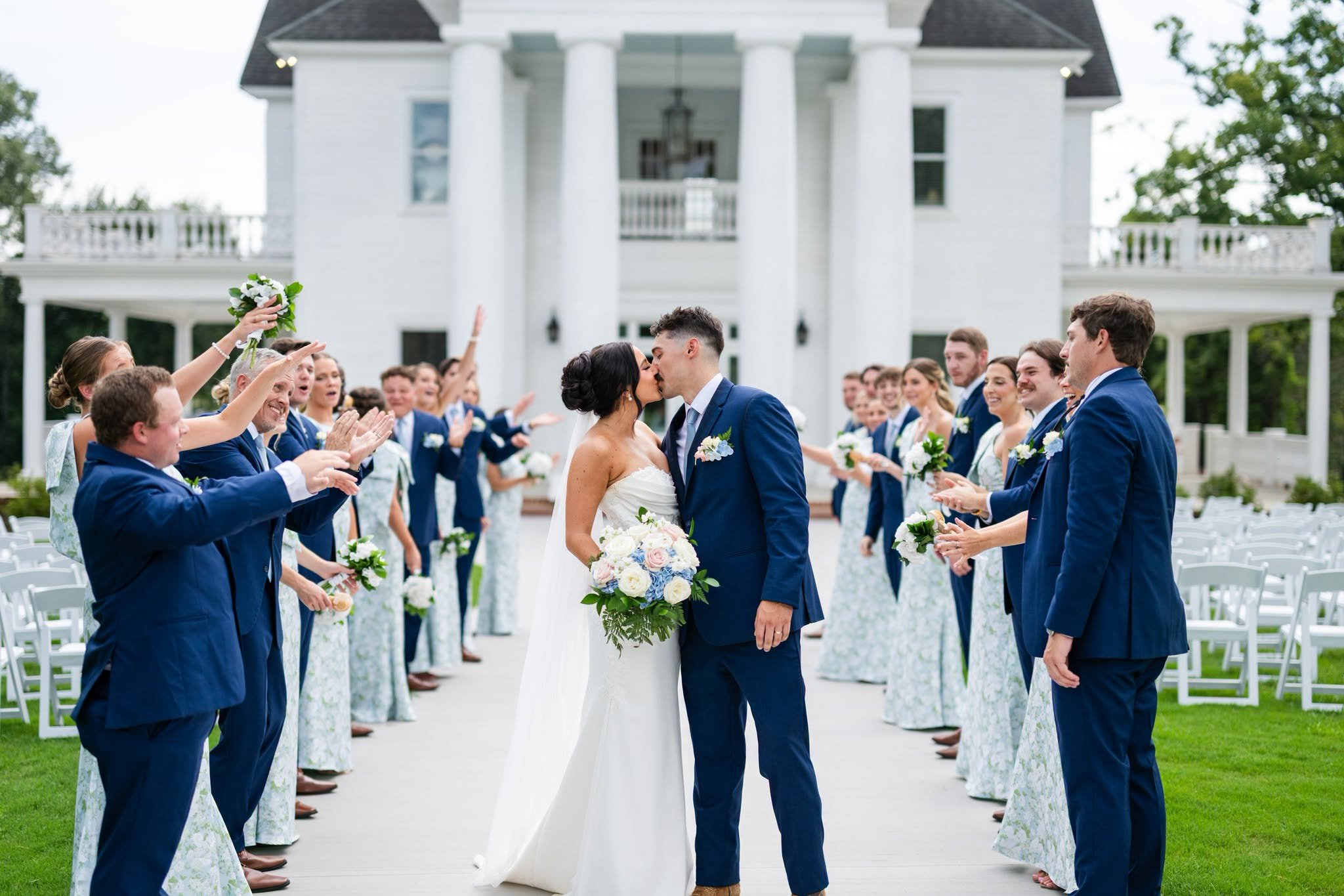 Bride and groom kissing as their wedding party cheers along the aisle in front of the white columned house at Heather Hill wedding venue.