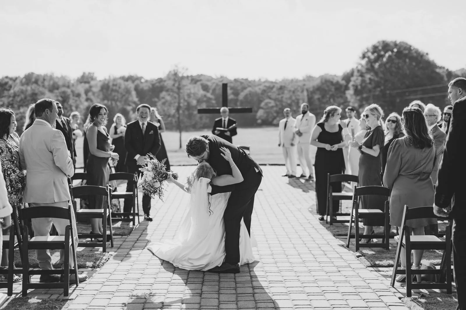 Black and white photo of a groom dipping and kissing his bride in the outdoor ceremony aisle at The Barn at Sitton Hill as guests smile around them.