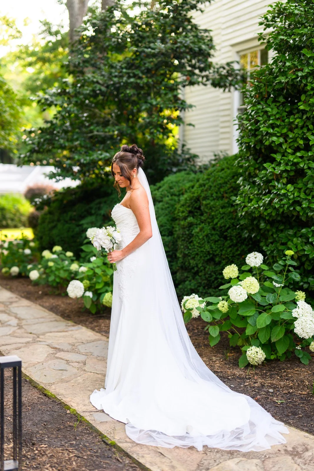 Bride holding a white bouquet on a stone garden path beside blooming hydrangeas at Duncan Estate in Spartanburg, SC.