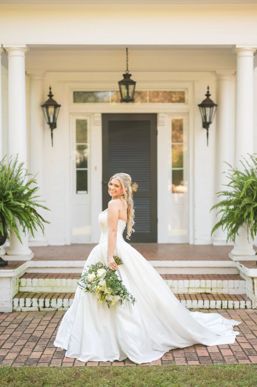 Bride smiling over her shoulder while holding a white and green bouquet outside the white house at The Barn at Sitton Hill, her gown trailing across the brick walkway.