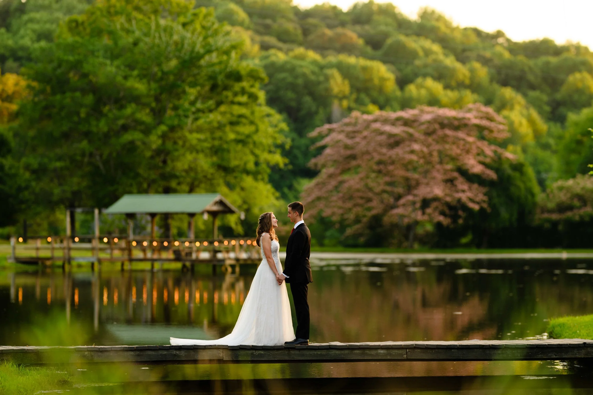 Bride and groom holding hands on a wooden bridge over the pond at South wind Ranch, with flowering trees and a pavilion reflected in the water behind them.