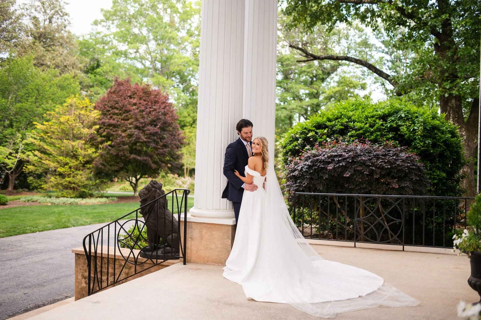 Bride and groom embracing beside a white column on Duncan Estate’s front porch, with the bride’s train spread across the porch in Spartanburg, SC.