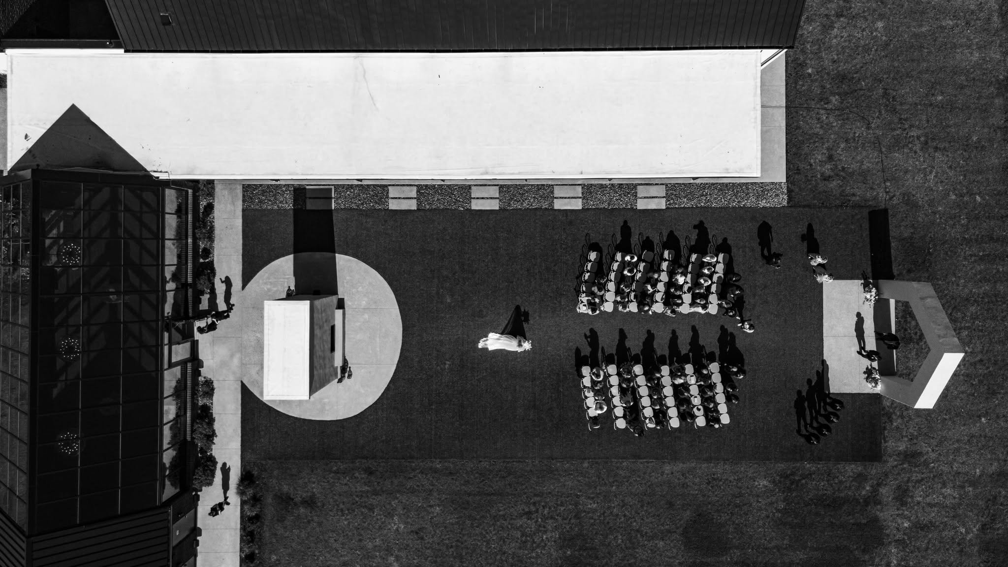 Black-and-white aerial view of a bride walking down the outdoor ceremony aisle at The Lumen House, with seated guests casting long shadows across the lawn.