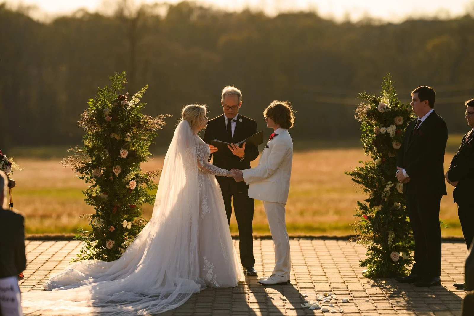 Bride in a lace gown and groom in a white suit holding hands during their outdoor ceremony at The Barn at Sitton Hill, framed by flowers and golden evening light.