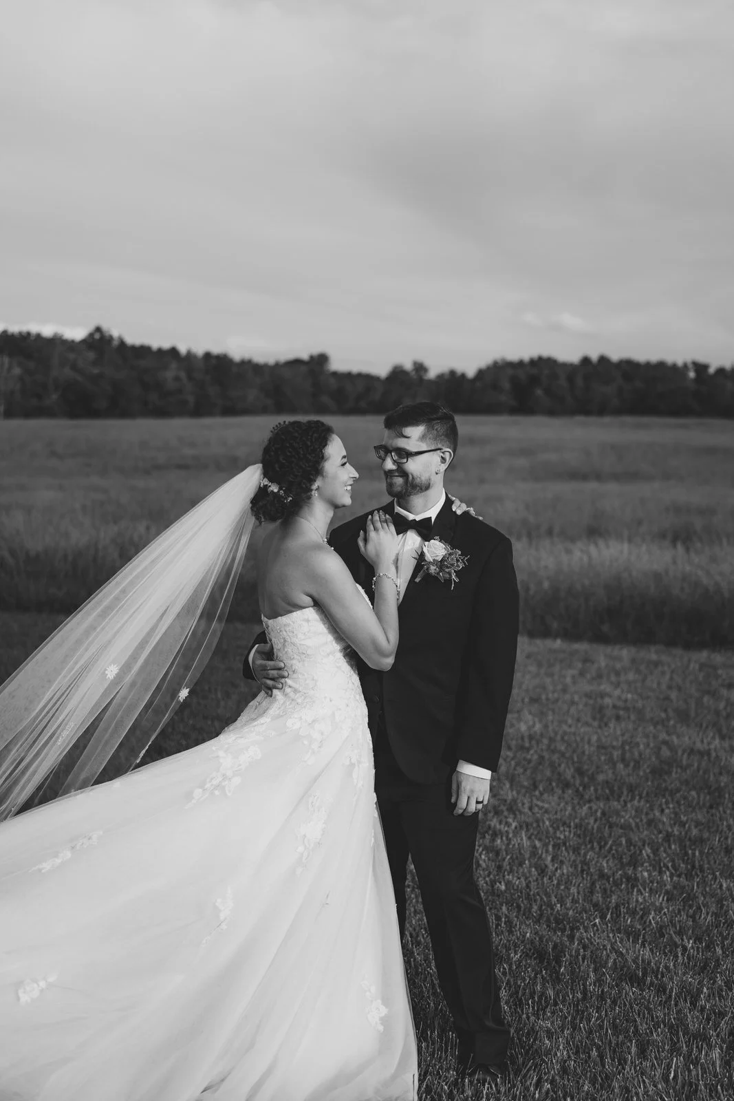 Black and white portrait of a bride and groom smiling at each other in MeadowView’s open field, with her veil and dress flowing to the left.
