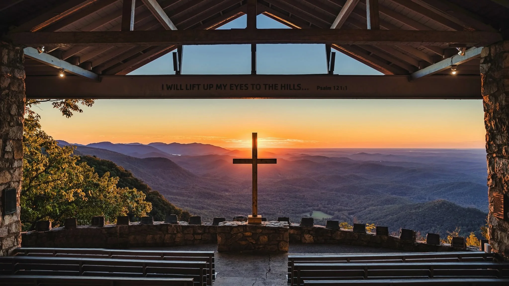 Sunrise behind the wooden cross at Pretty Place Chapel, overlooking layered mountains with empty pews in the foreground.