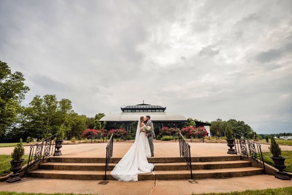 Bride and groom kissing on the outdoor steps at Edinburgh West, with her gown trailing down the stairs and the glass conservatory and flowering gardens behind them.