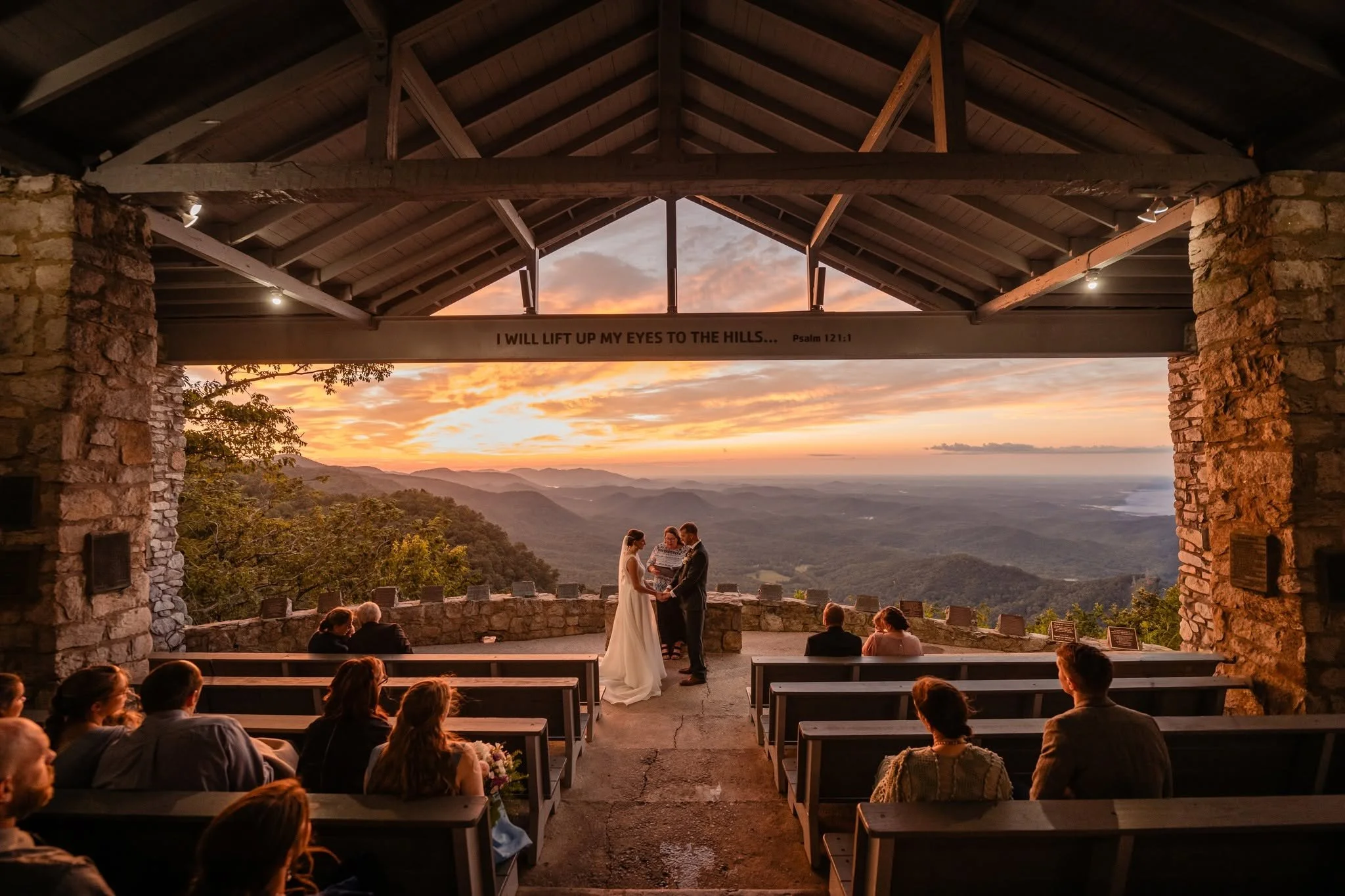 Bride and groom holding hands during a wedding ceremony at Pretty Place Chapel, with guests seated beneath wooden beams and an orange sunrise over the mountains.