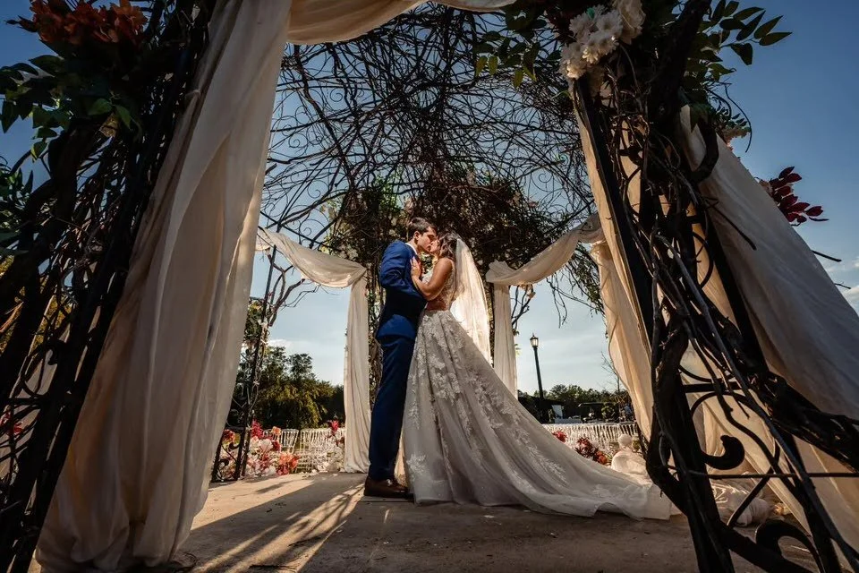 Bride and groom kissing beneath Edinburgh West’s ornate iron gazebo, framed by white draping and flowers, with her lace train trailing behind her.