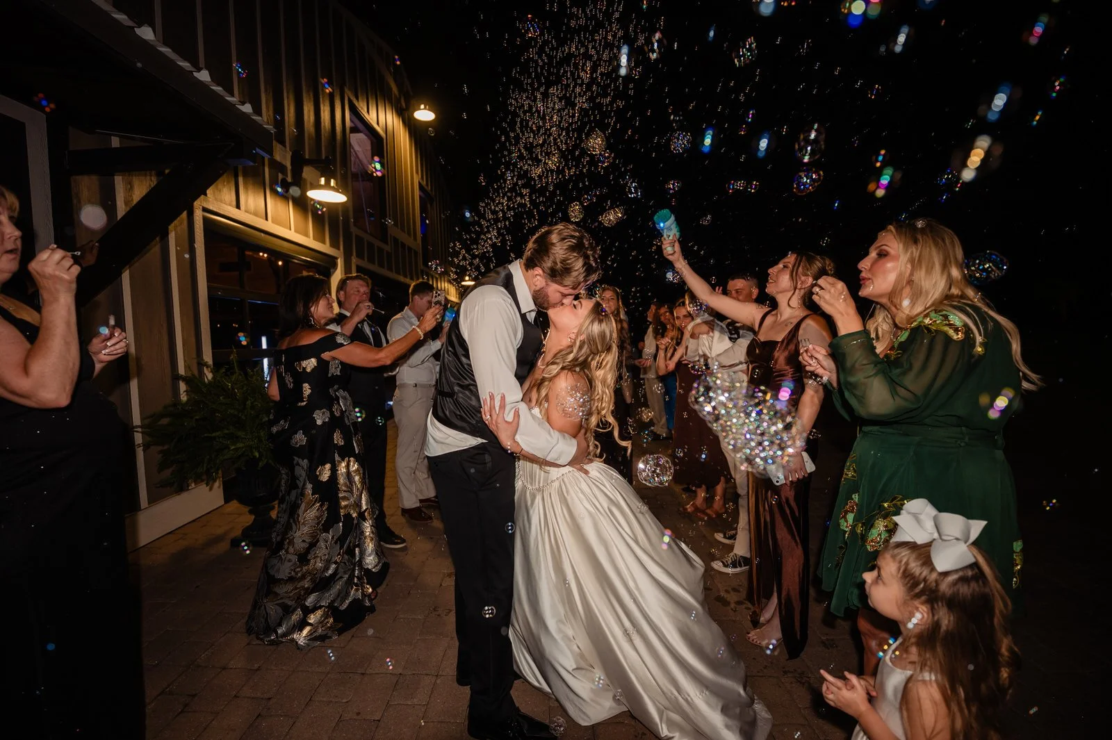 Bride and groom kissing during their nighttime bubble send-off outside The Barn at Sitton Hill as family and friends surround them blowing bubbles.