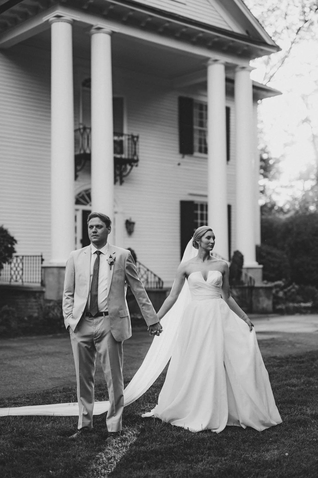 Black and white portrait of a bride and groom holding hands on the lawn in front of Duncan Estate’s grand columns in Spartanburg, SC.