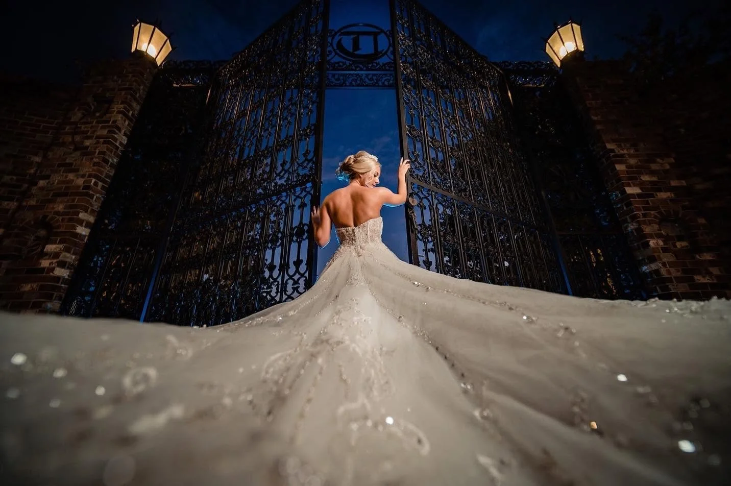 “Bride standing between ornate iron gates at Edinburgh West at night, with her wedding gown’s long train filling the foreground and lanterns glowing above.”