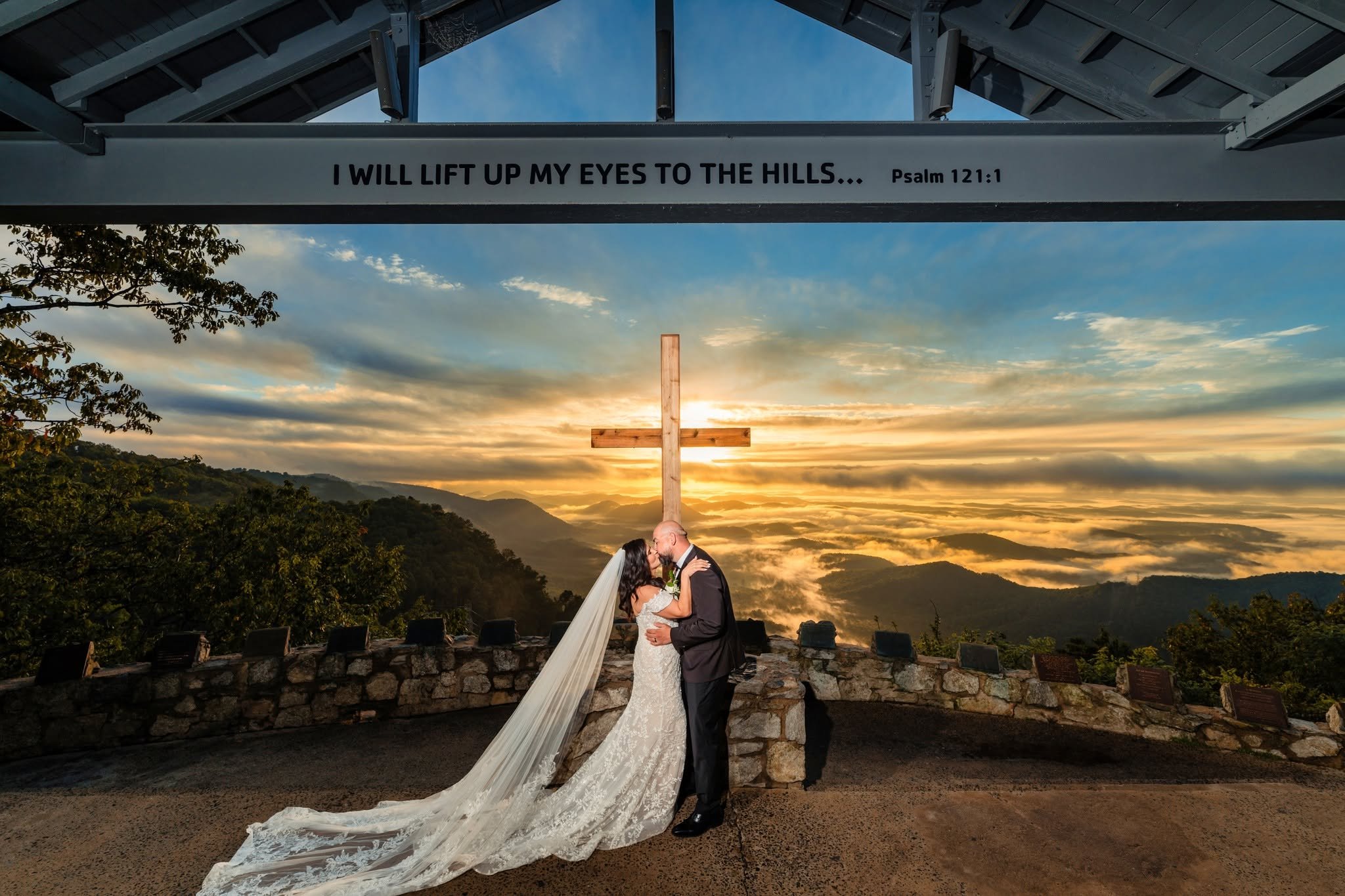 Bride and groom kissing beneath the wooden cross at Pretty Place Chapel, with her long veil trailing across the floor and golden sunrise over misty mountains.