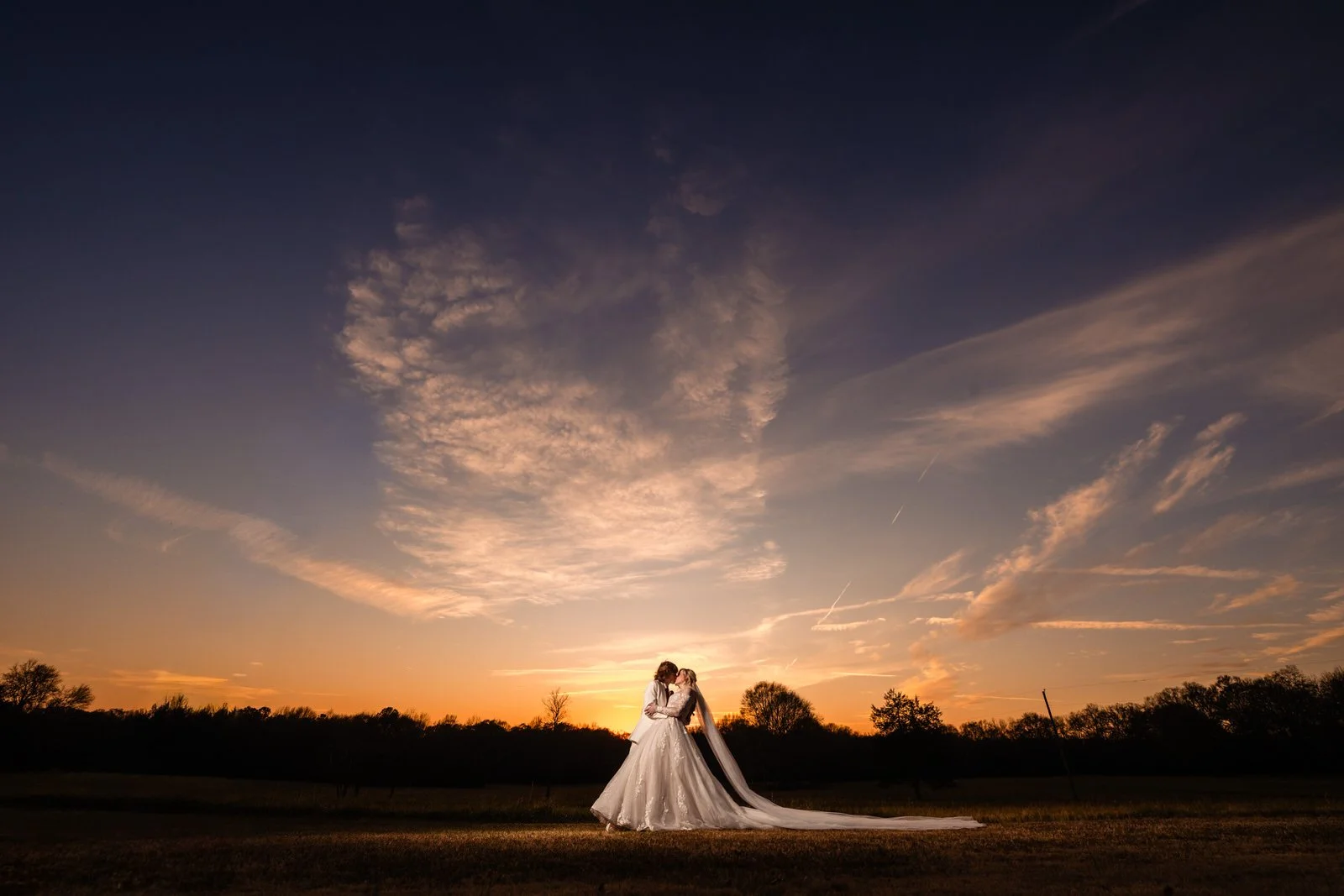Bride and groom embracing in a field at The Barn at Sitton Hill beneath a sweeping sunset sky, with her long veil trailing across the grass.