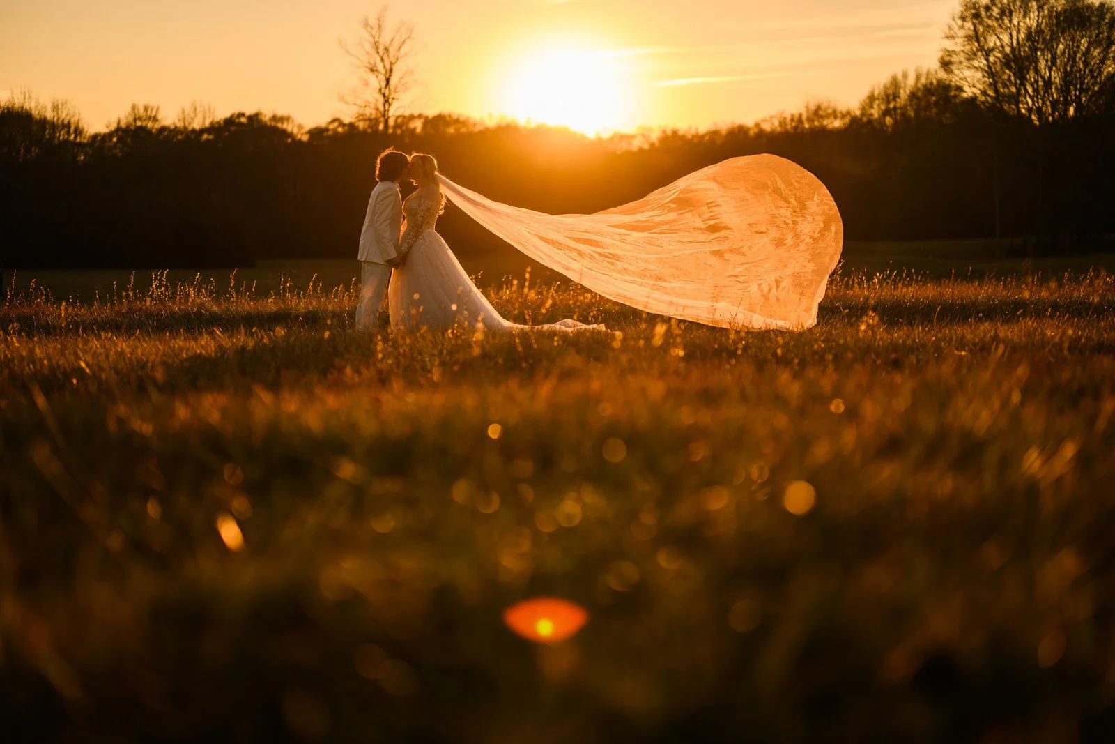 Bride and groom kissing in a field at The Barn at Sitton Hill as the setting sun illuminates her long veil billowing behind her.