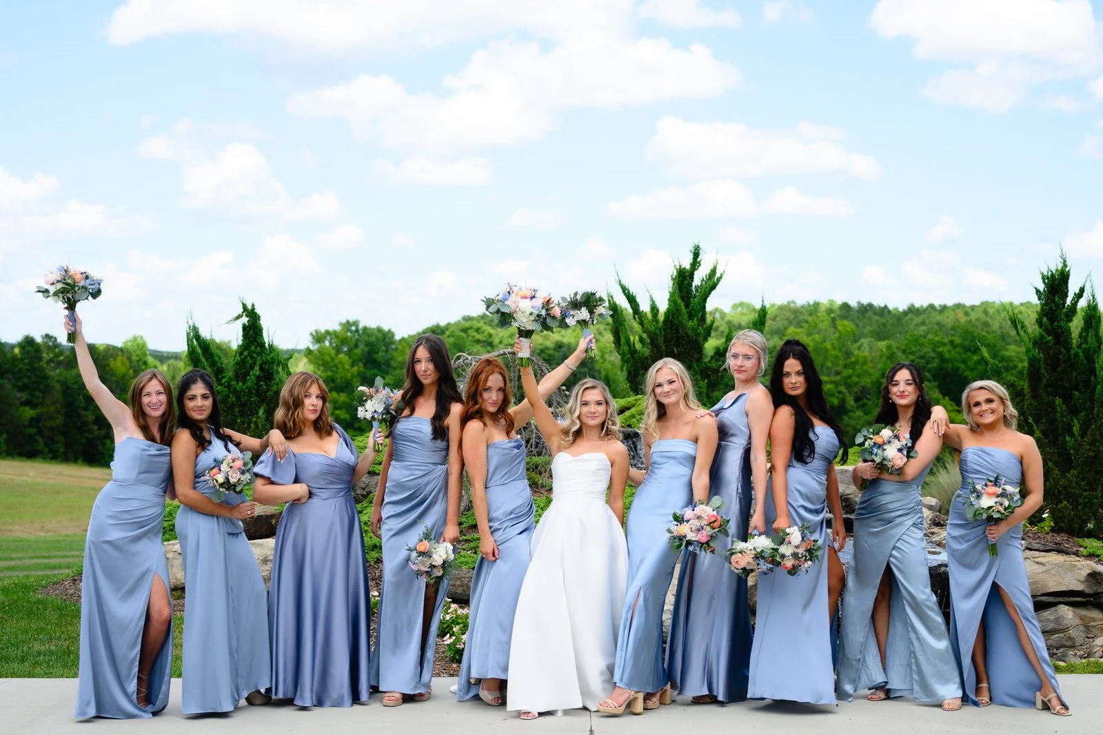Bride surrounded by bridesmaids in blue dresses holding pastel bouquets beside MeadowView’s landscaped rock garden under a blue sky.