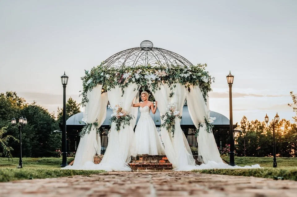 Bride standing beneath Edinburgh West’s ornate iron gazebo, surrounded by white draping, flowers, and greenery in the evening light.
