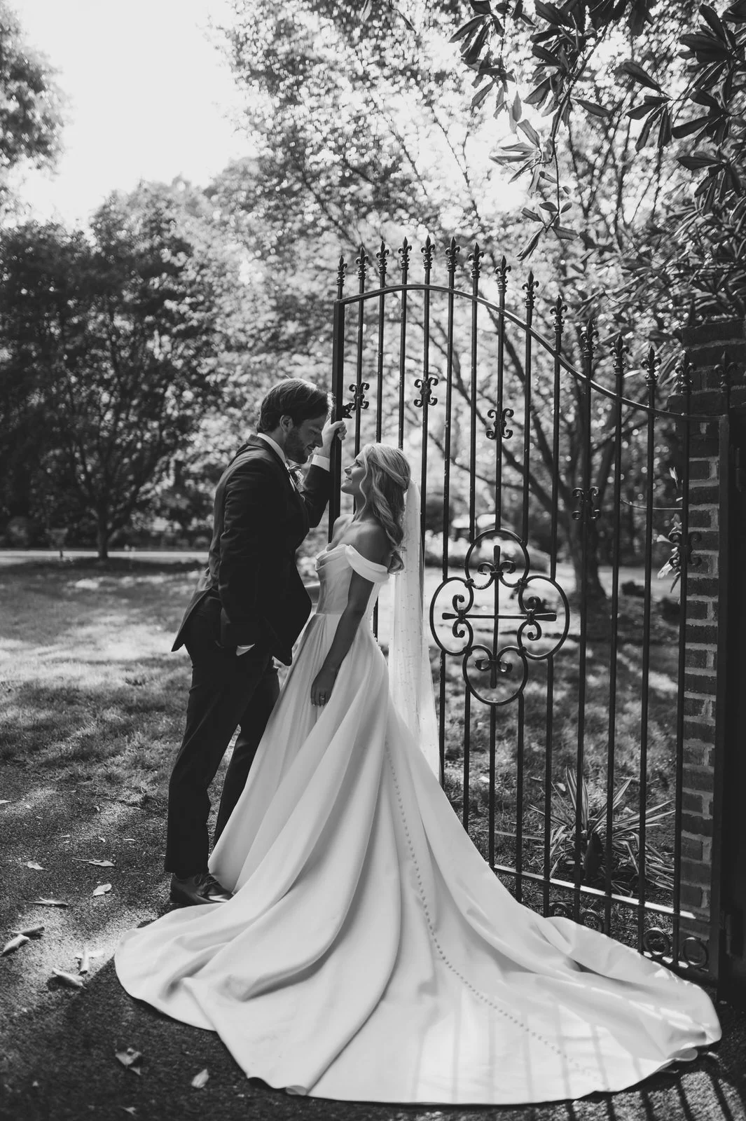 Black and white portrait of a bride and groom beside an ornate iron gate at Duncan Estate in Spartanburg, SC, with the bride’s train spread across the ground.