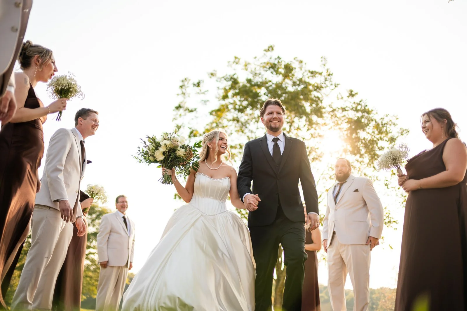 Smiling bride and groom walking hand in hand between bridesmaids in brown dresses and groomsmen in light suits at The Barn at Sitton Hill, with sunlight streaming through the trees.