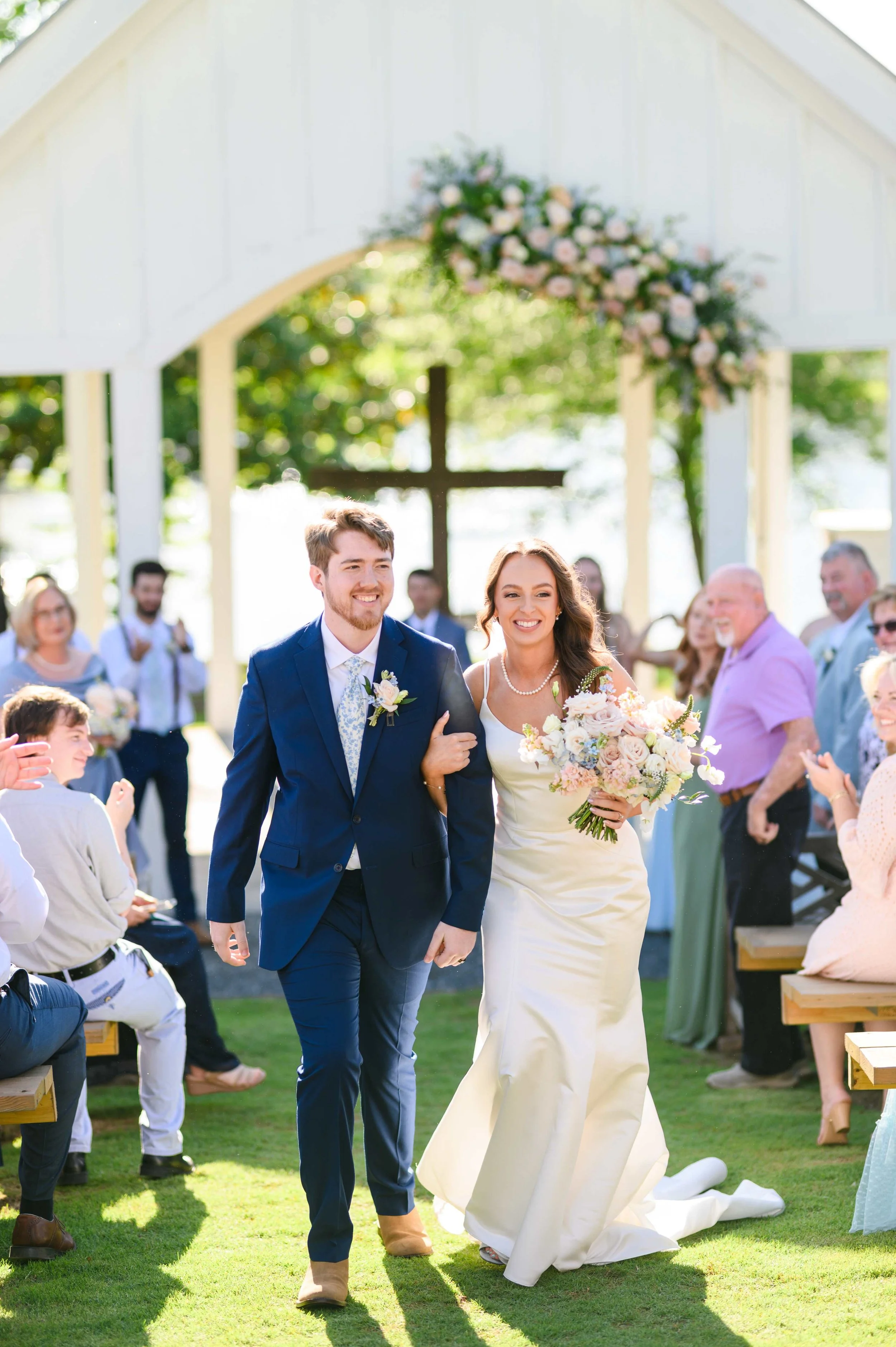 Smiling bride and groom walking arm in arm down the aisle at South Wind Ranch as guests applaud, with the flower-decorated white chapel behind them.