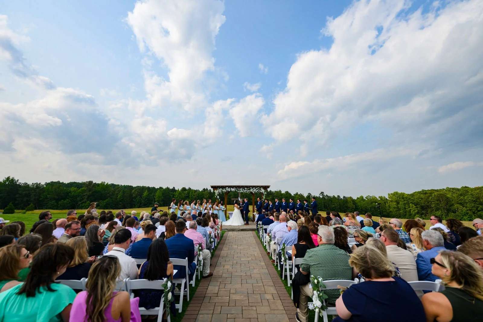 Wide view down the paved aisle at MeadowView’s outdoor wedding ceremony, with guests in white chairs and the couple beneath a floral pergola under a blue, cloud-filled sky.