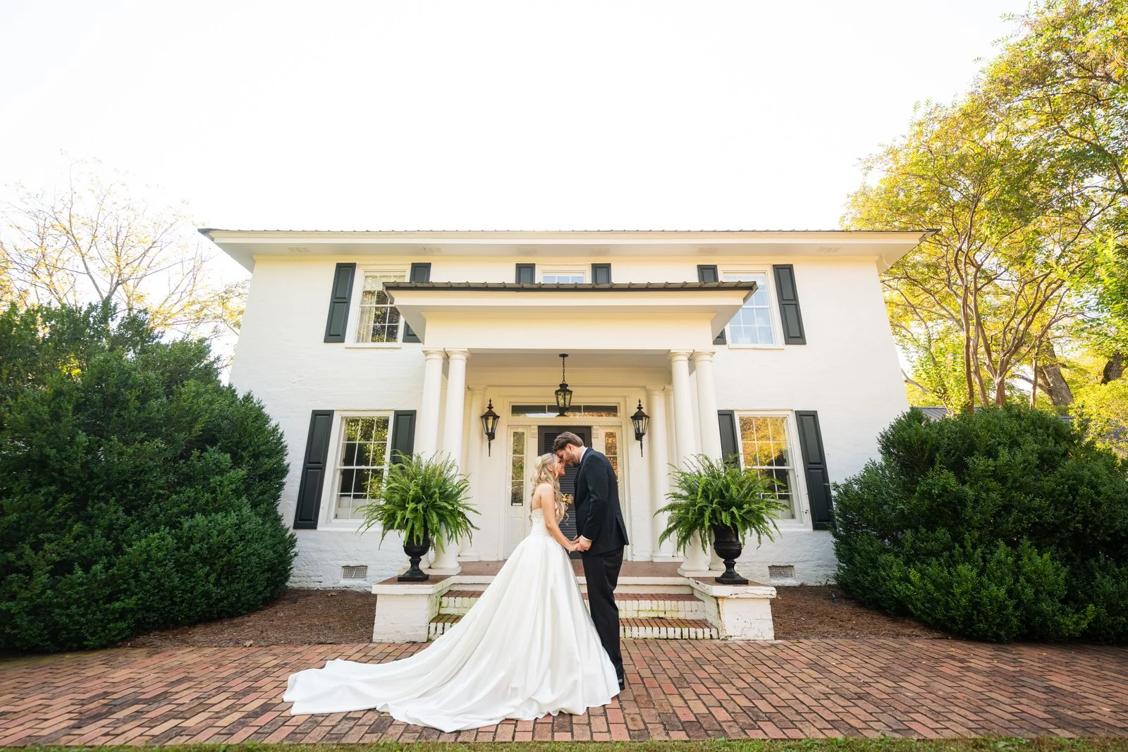 Bride and groom holding hands with their foreheads together outside the white house at The Barn at Sitton Hill, her long train spread across the brick walkway.