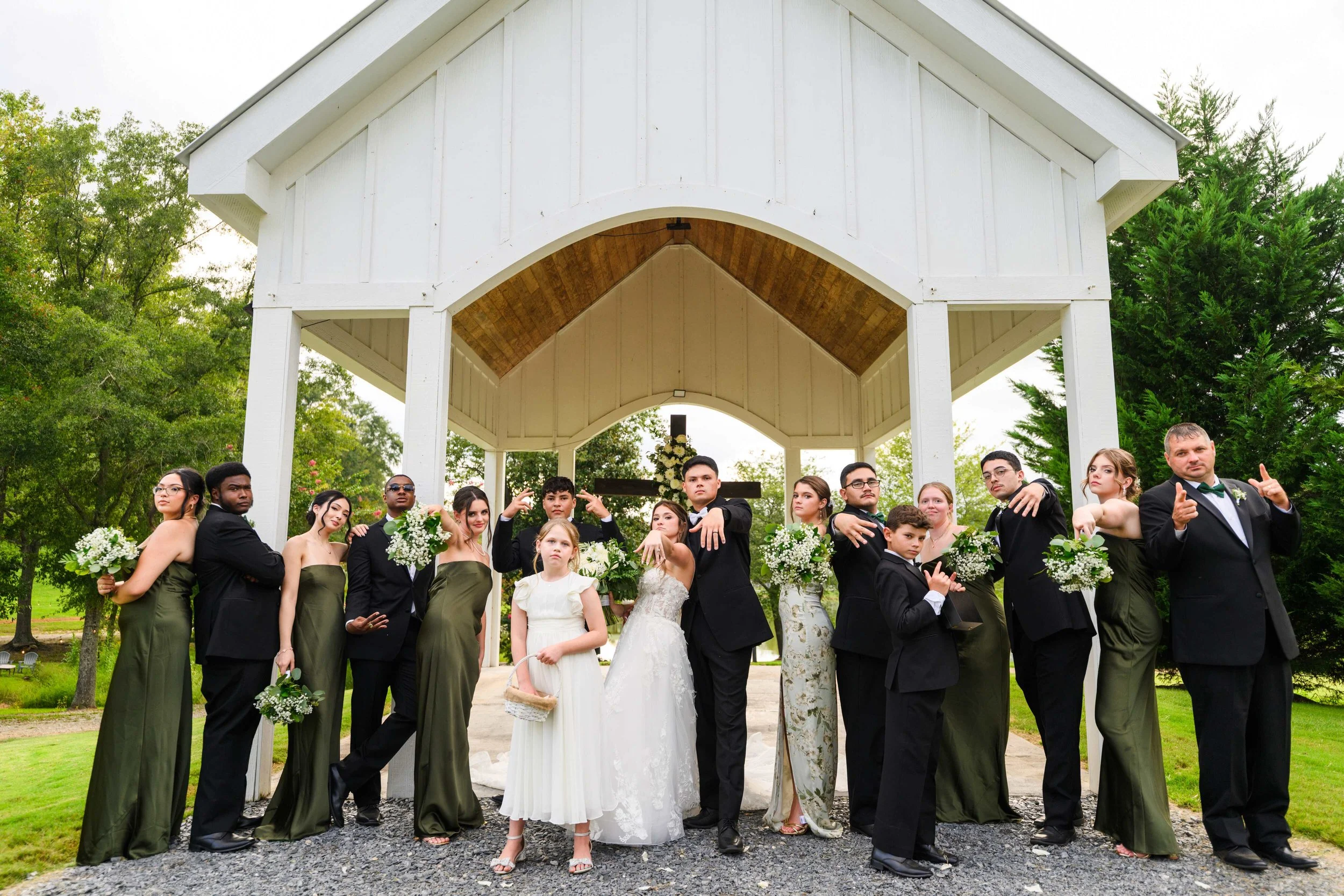 Bride, groom, and wedding party striking playful poses in front of the white open-air chapel at South Wind Ranch, with green bridesmaid dresses and black suits.