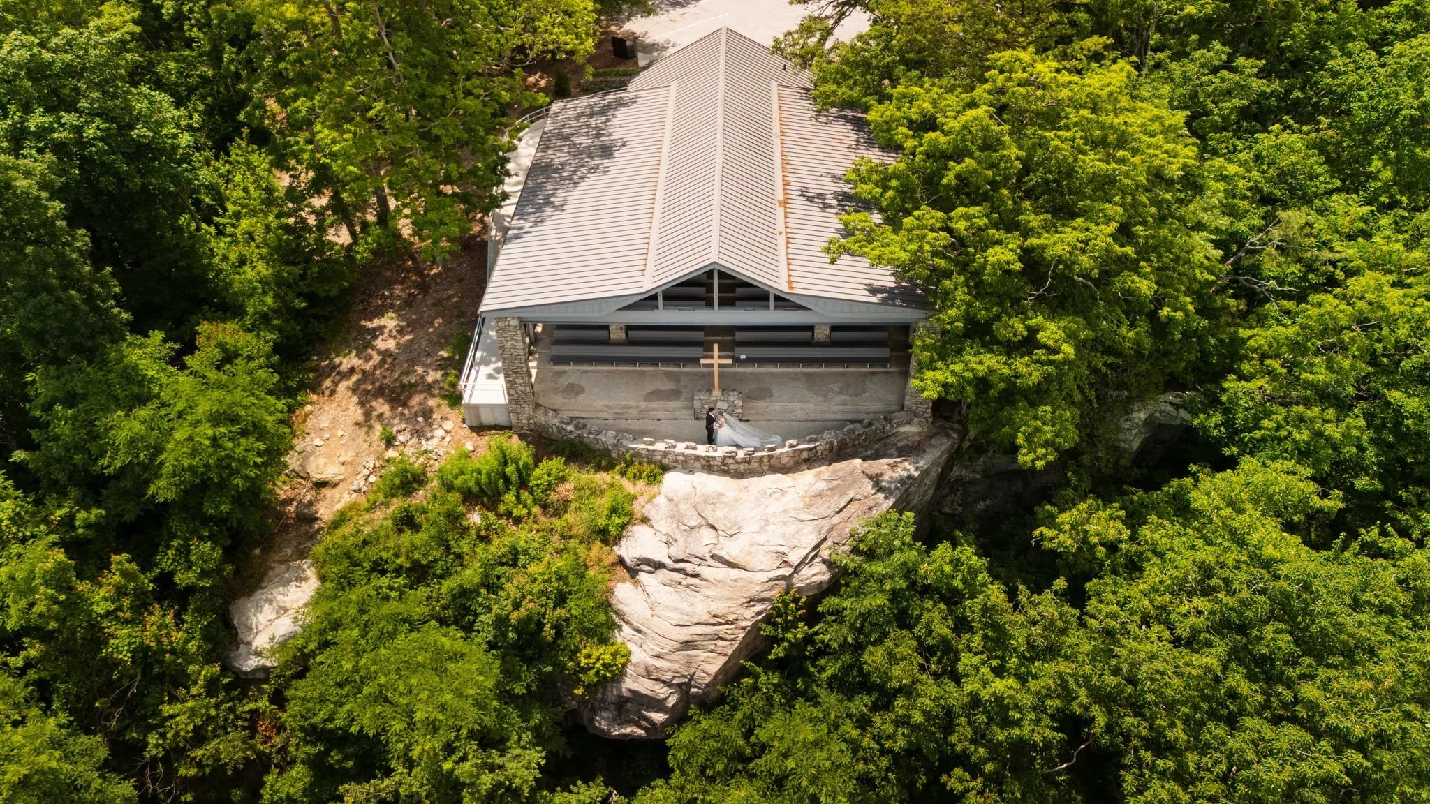 Aerial view of a bride and groom beneath the cross at Pretty Place Chapel, with the chapel perched above a rocky ledge surrounded by green forest.