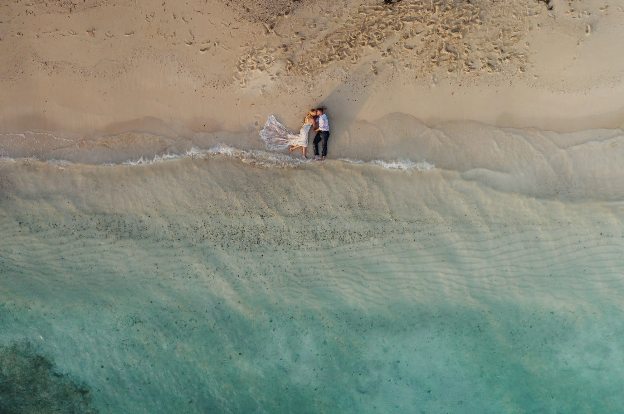 Overhead aerial view of a bride and groom lying together at the water’s edge on a sandy beach in Jamaica, with gentle waves and turquoise water