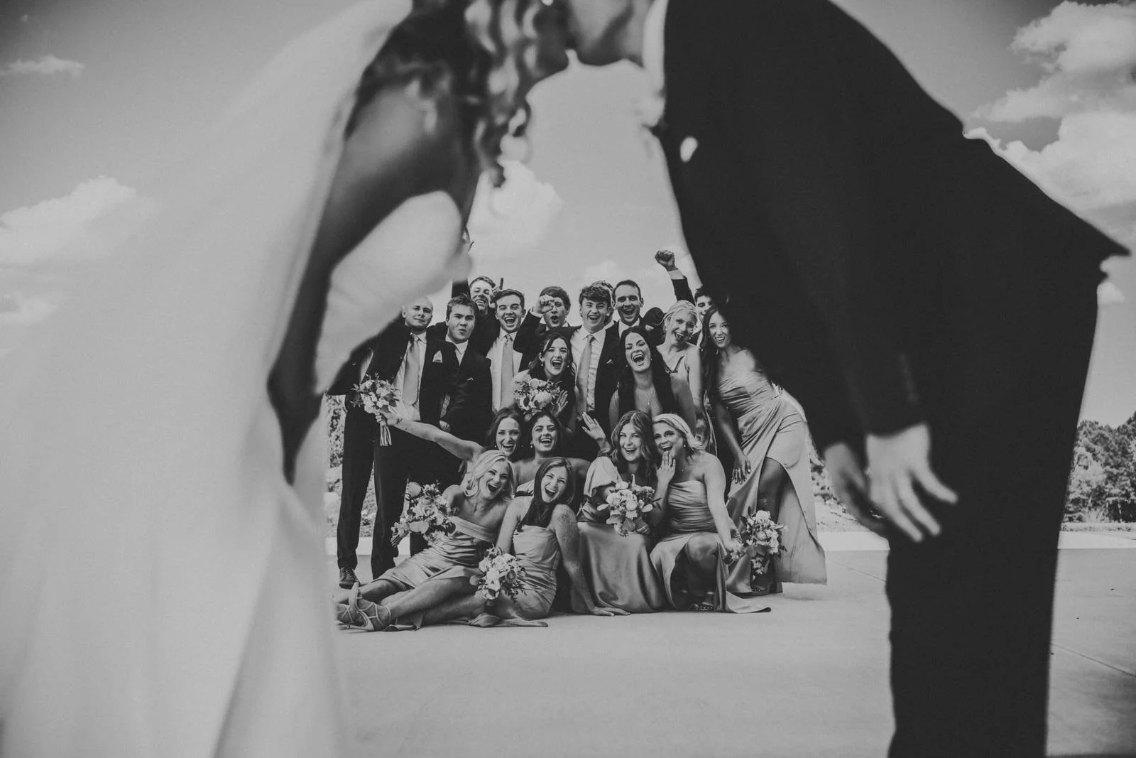 Black and white photo of a bride and groom kissing in the foreground while their wedding party laughs and cheers behind them at MeadowView.